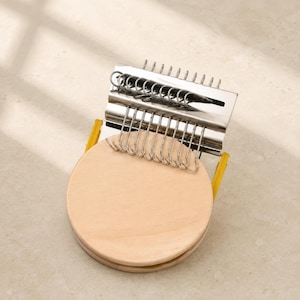 May include: A darning loom tool featuring a round, light-coloured wooden base and a silver-toned metal top component. The metal section includes a series of vertical hooks and a sliding mechanism. Two yellow plastic side supports connect the two parts.