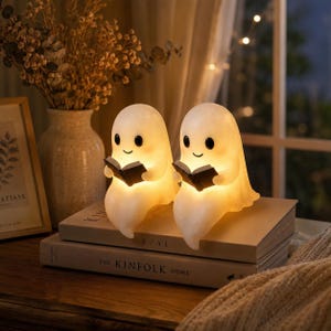 May include: Two small, glowing, off-white ghost-shaped lamps, each featuring a smooth, rounded head, a simple smiling face with black eyes, and holding a small, open black book. The lamps emit a warm, soft light from within their translucent bodies.