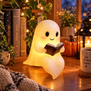 May include: A glowing, off-white ghost-shaped lamp with a smooth texture, black oval eyes, and a small smile. The ghost figure is holding a small, dark-colored open book in its hands. The lamp emits a warm, soft light, illuminating its form.