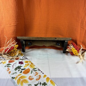Op de afbeelding: Een donkerbruine houten wandplank met een plat bovenoppervlak en twee kleine ronde haken aan de onderkant. De plank is geplaatst tegen een oranje stoffen achtergrond, met decoratieve gedroogde maïskolven en een witte loper met herfstbladeren.