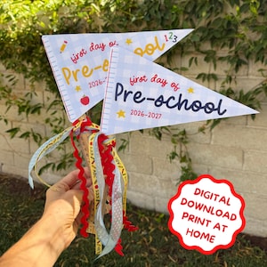 May include: Two triangular pennants featuring a light blue gingham pattern, yellow stars, and the text "first day of Pre-school 2026-2027". The pennants are attached to wooden sticks decorated with colourful ribbons in red, yellow, and light blue.