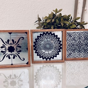 May include: Three square wooden frames with black and white geometric designs on a silver background. The frames are arranged in a row on a white surface.