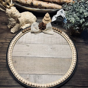 May include: A round wooden tray with a whitewashed woodgrain finish. The tray is decorated with a white bead border and a burlap bow with a small pine cone in the center. The tray is sitting on a dark wood surface. #1