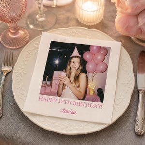 Op de afbeelding: Een vierkant wit papieren servet met een afgedrukte foto van een persoon die een roze feesthoedje draagt en een cupcake vasthoudt. Onder de afbeelding staat de tekst HAPPY 18TH BIRTHDAY! en de naam Louisa in een roze lettertype.
