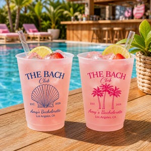 May include: Two frosted plastic cups filled with a pink beverage, ice, and fruit slices. The left cup features a blue seashell graphic and text reading THE BACH Club EST. 2026 Amy's Bachelorette Los Angeles, CA. The right cup has a pink palm tree graphic.