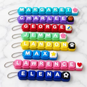 May include: Eight colorful silicone keychains featuring white block letters spelling names including Margaret, Charlee, George, Jemma, Landon, Max, Lawrence, and Elena, each finished with a small charm like a flower, ball, or heart.
