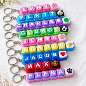 May include: A collection of nine colorful silicone keychains, each featuring block letters spelling names including EMMY, MERTT, CHARLEE, GEORGE, JEMMA, LANDON, JACOB, MAX, and ELENA, with attached charms like flowers, sports balls, stars, hearts, and paw prints.