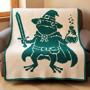 Puede incluir: Una manta de ganchillo de color crema con un gran gráfico pixelado de color verde oscuro de una rana con sombrero de mago y capa. La rana sostiene una espada en una mano y un frasco de poción burbujeante en la otra, con un cinturón y bolsa.