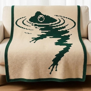 May include: A rectangular crochet blanket featuring a cream-colored base with a dark green graphic design of a frog swimming in water. The frog is depicted with ripples surrounding its head, and the blanket has a solid dark green border.