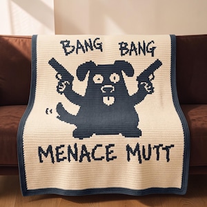 May include: A cream-colored crochet blanket featuring a dark blue graphic of a dog holding two pistols. The text BANG BANG is positioned above the dog, and the text MENACE MUTT is below. The blanket has a dark blue border around the edges.