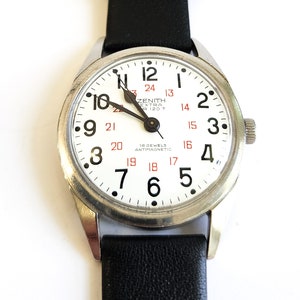 May include: A silver Zenith Extra 120 T wristwatch with a white face and black leather band. The watch has black Arabic numerals and red markings for the hours. The watch face also includes the text "Zenith Extra 120 T" and "18 Jewels Antimagnetic".