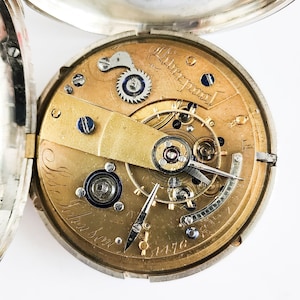 Könnte beinhalten: Nahaufnahme des Innenlebens einer silbernen Taschenuhr. Das Zifferblatt der Uhr ist goldfarben und trägt die Inschrift "Liverpool" und "J.B. Johnson".