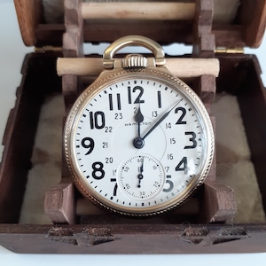 May include: A gold pocket watch with a white face and black hands. The watch is in a wooden box with a velvet lining. The watch face has black Roman numeral hour markers and black Arabic numeral minute markers. The watch has a small second hand dial at the bottom of the face. The watch is labelled "Hamilton" at the top of the face.