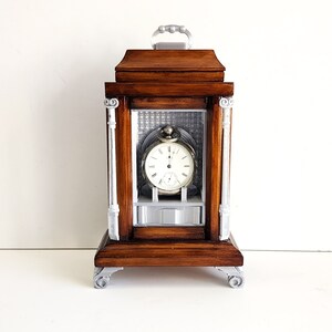 May include: A wooden clock with a silver face and hands, enclosed in a decorative wooden frame with silver accents. The clock has a silver handle on top and sits on four silver feet.