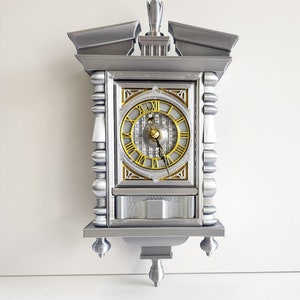 May include: A silver-coloured clock with a gold face and black Roman numeral markings. The clock has a decorative design with a pointed top and ornate details.