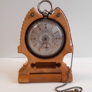 May include: A silver pocket watch with a white face and black Roman numeral hour markers is displayed in a wooden stand. The watch has a chain attached to it and a small screwdriver is lying on the table.