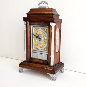 May include: A wooden clock with a gold face and silver accents. The clock has a decorative handle on top and sits on four silver feet.