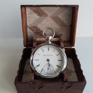 May include: A silver pocket watch with a white face and black numerals, displayed in an open wooden box with a patterned interior. The watch has a small seconds dial and the text "Elgin Natl. Watch Co."
