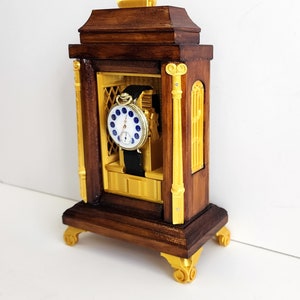 Puede incluir: Un soporte de reloj de madera con detalles dorados y un reloj de bolsillo en su interior. El reloj tiene una esfera blanca con números romanos azules y una correa de cuero negra.