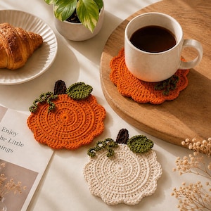 May include: Three round crochet coasters shaped like pumpkins, featuring scalloped edges, a dark brown stem, a green leaf, and curly green vines. Two coasters are vibrant orange, and one is off-white. One orange coaster supports a white ceramic mug.