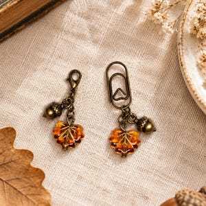 May include: Two bronze-toned metal accessories featuring amber-colored glass leaf charms and small acorn charms. One accessory is a lobster clasp charm, and the other is a paperclip-style marker, both showcasing detailed autumn-inspired designs.
