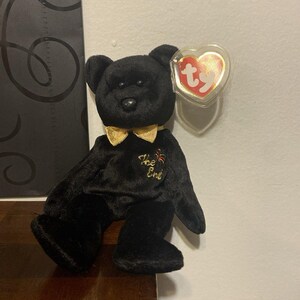 Könnte beinhalten: Ein schwarzer Plüschbär mit einer goldenen metallischen Fliege um den Hals und dem gestickten Text The End in Goldfaden auf der Brust. Der Bär hat ein herzförmiges rot-goldenes Ty-Etikett am linken Ohr, das mit einer durchsichtigen Kunststoffhülle geschützt ist.