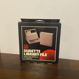 Peut inclure: Une boîte de rangement noire Acco Diskette Library File avec une poignée. Le devant présente le texte blanc Diskette Library File, Holds and Displays 15 5.25 inch diskettes, et un graphique d'un porte-disquette beige et une sphère rouge.