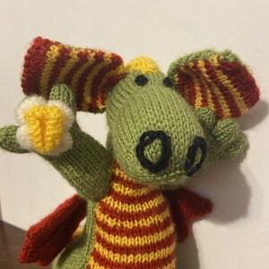 May include: A hand-knitted green dragon toy featuring a yellow and red striped belly, red and yellow striped ears, and black embroidered nostrils. The dragon holds a small white flower with a yellow center in one of its knitted green claws.