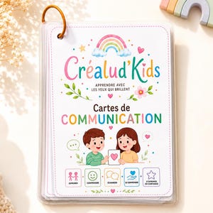 Puede incluir: Un juego de tarjetas de comunicación en una anilla, con el título Créalud'Kids, Apprendre avec les yeux qui brillent y Cartes de COMMUNICATION. La portada muestra un arcoíris, detalles florales y dos niños sosteniendo una tarjeta de corazón.