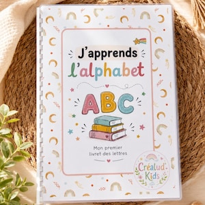 Peut inclure: Un livret éducatif à reliure spirale intitulé J'apprends l'alphabet avec une couverture blanche ornée d'illustrations colorées d'arcs-en-ciel, d'étoiles et de lunes. Le centre affiche les lettres ABC au-dessus de trois livres et le texte Mon premier livret des lettres.