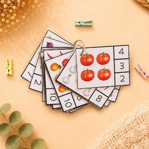 Peut inclure: Un ensemble de cartes éducatives pour apprendre à compter, reliées par un anneau métallique. Chaque carte présente des illustrations de fruits ou légumes et un chiffre. La carte du dessus montre quatre tomates rouges et les chiffres 4, 3 et 2.