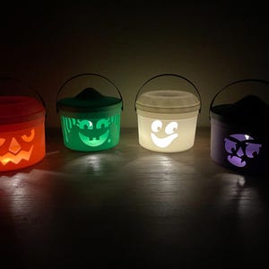 May include: Four cylindrical plastic buckets with lids and handles, each featuring a unique cutout face design. The buckets are illuminated from within, glowing in red, green, white, and purple, casting light onto the surface below them.
