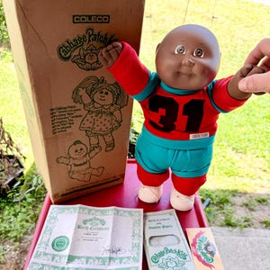 May include: A Cabbage Patch Kids doll with dark skin, wearing a red and teal athletic outfit with the number 31 on the chest. The doll is accompanied by its original cardboard box, a birth certificate, adoption papers, and small paper inserts.