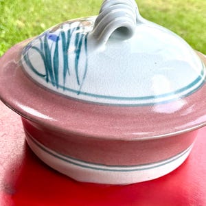 May include: A round ceramic lidded casserole dish featuring a glossy white base with a wide dusty rose band and a matching lid. The lid has a hand-painted floral design in green and blue with a prominent, thick, looped handle at the center.