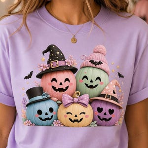 May include: A light purple t-shirt featuring a graphic of five colourful pumpkins with faces. The pumpkins wear accessories including a black witch hat, a pink knit beanie with a bat, a blue top hat, a purple polka dot bow, and a pink witch hat.