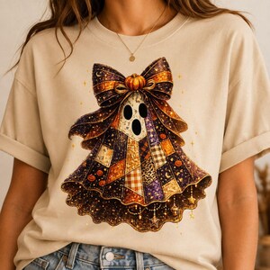 May include: A beige t-shirt featuring a graphic of a ghost wearing a large, patterned bow on its head. The ghost's body is composed of various patchwork squares in shades of orange, purple, and black, decorated with small pumpkins and stars.