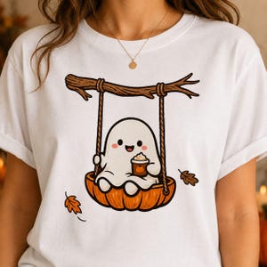 May include: A white t-shirt featuring a graphic of a small white ghost sitting on a swing made from a pumpkin. The ghost is holding a brown coffee cup with a pumpkin icon. A brown tree branch serves as the swing support, with falling autumn leaves.