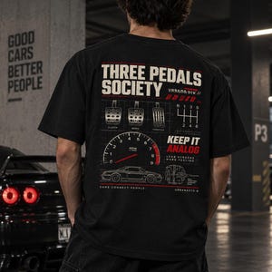 Op de afbeelding: Een zwart t-shirt met een witte en rode afbeelding op de achterkant. Het ontwerp bevat de tekst "THREE PEDALS SOCIETY", "KEEP IT ANALOG" en "CARS CONNECT PEOPLE", samen met illustraties van autopedalen, een versnellingspatroon en een toerenteller.