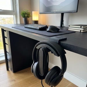 Puede incluir: Un soporte para auriculares negro impreso en 3D montado en el borde de un escritorio. El soporte cuenta con un brazo curvo que sostiene unos auriculares negros de diadema con un cable en espiral. La pieza tiene una superficie plana superior.