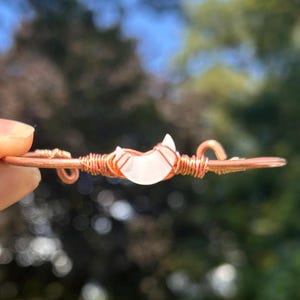 Könnte beinhalten: Ein Armreif aus Kupferdraht mit einem zentralen, halbmondförmigen Stein in blasser, durchscheinender weißer Farbe. Der Metalldraht ist fest um die Basis des Steins gewickelt und erstreckt sich nach außen, um das Armband zu bilden.