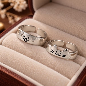 Puede incluir: Dos anillos abiertos de tono plateado con motivos de animales. Un anillo muestra una cara de perro con orejas caídas y un pequeño hocico, mientras que el otro representa una cara de gato con orejas puntiagudas y bigotes. Acabado pulido.