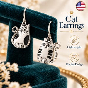 May include: A pair of silver-toned drop earrings featuring a cat design with black enamel accents. The cat figures have wide eyes, whiskers, and curled tails. The text on the right side reads Cat Earrings, Lightweight, and Playful Design.