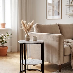 Peut inclure: Une table d'appoint ronde avec un plateau en faux marbre blanc et une étagère inférieure, soutenue par un cadre en métal noir à barres verticales. Un petit vase en céramique bicolore avec des herbes séchées et une tasse reposent dessus.