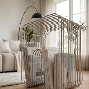 Peut inclure: Un porte-revues en métal beige clair composé d'une série de tiges métalliques verticales parallèles formant une structure arquée. Le support contient plusieurs livres et magazines aux couvertures blanches, dont Design as Art et Kinfolk.