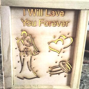 May include: A square wooden box frame featuring a light-coloured wood finish. The front panel displays the laser-cut text I Will Love You Forever in gold, alongside gold-toned illustrations of a couple, two overlapping hearts, and two rings.