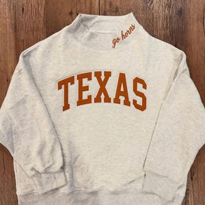 May include: A light gray heathered sweatshirt with a mock neck collar. The word TEXAS is printed in large, arched, burnt orange block letters across the chest. The phrase go horns is embroidered in cursive burnt orange thread on the collar.