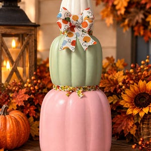 May include: A vertical stack of three decorative pumpkins in white, sage green, and soft pink. The top white pumpkin features a bow with a colorful pumpkin print, while a thin ribbon with a leaf pattern separates the green and pink pumpkins.