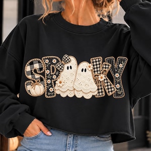 May include: A black long-sleeve sweatshirt featuring the word SPOOKY in a large graphic print. The letters are decorated with a black and white buffalo plaid pattern, white daisies, a small pumpkin, and two white ghosts with eyelet lace details.