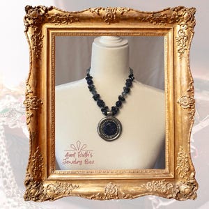 May include: A necklace featuring a large, circular, dark blue pendant with a silver-toned border, suspended from a strand of dark, faceted beads. The text "Aunt Ruth's Jewelry Box" is printed in the lower left corner of the display area.
