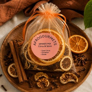 Peut inclure: Un sachet en organza orange transparent à cordon contenant un mélange à mijoter composé de tranches d'agrumes séchées, de bâtons de cannelle et de clous de girofle. Une étiquette rose indique Afrogourmets Simmer Mix Citrus & Spice.